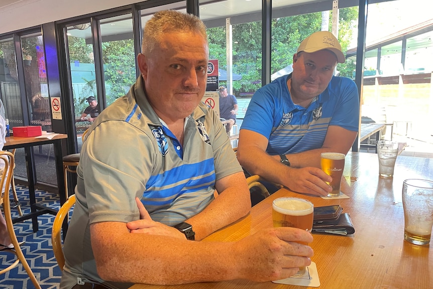 Two men sit at a table in a pub holding beers.