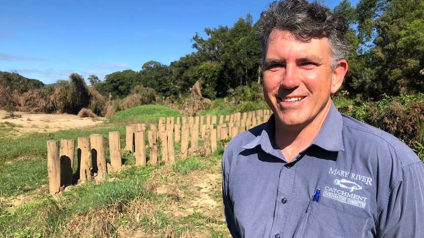 A man smiles at the camera with rows of posts driven into the riverbank behind him.