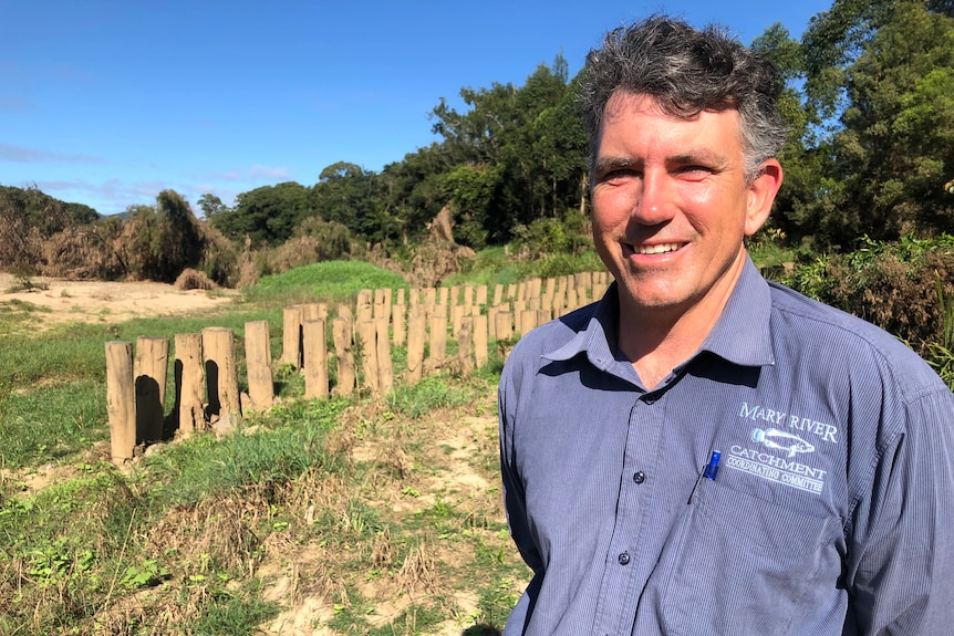 A man smiles at the camera with rows of posts driven into the riverbank behind him.