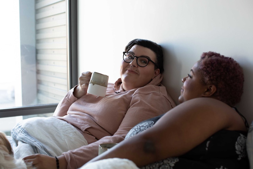 Two women in bed smile at each other, drinking coffee.