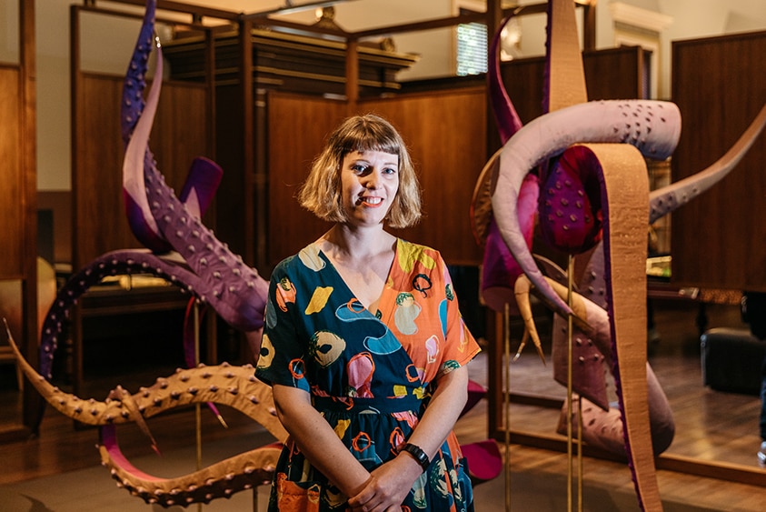 A smiling woman with blonde bob and colourful dress stands in front of sculpture with octopus tentacles indoors.