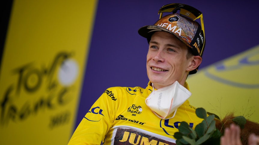 A rider in a yellow jersey smiles as he stands on the podium carrying a bouquet after a Tour de France stage. 