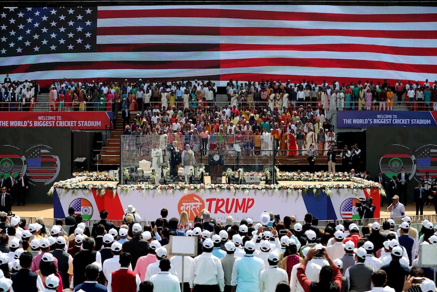 Donald Trump, Melania Trump and Narendra Modi stand on the stage in front of a large screen displaying the American flag.