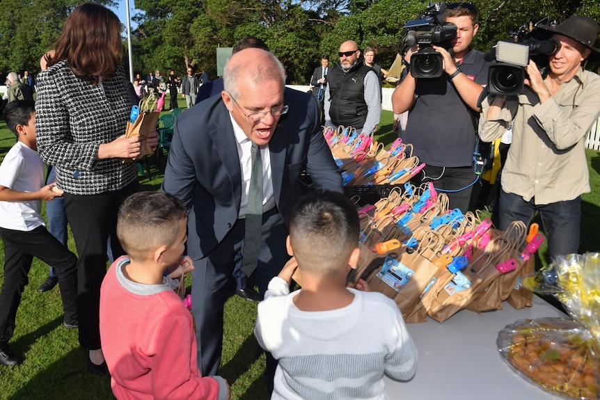 A man opens his mouth wide as he hands out bags to two kids, surrounded by media