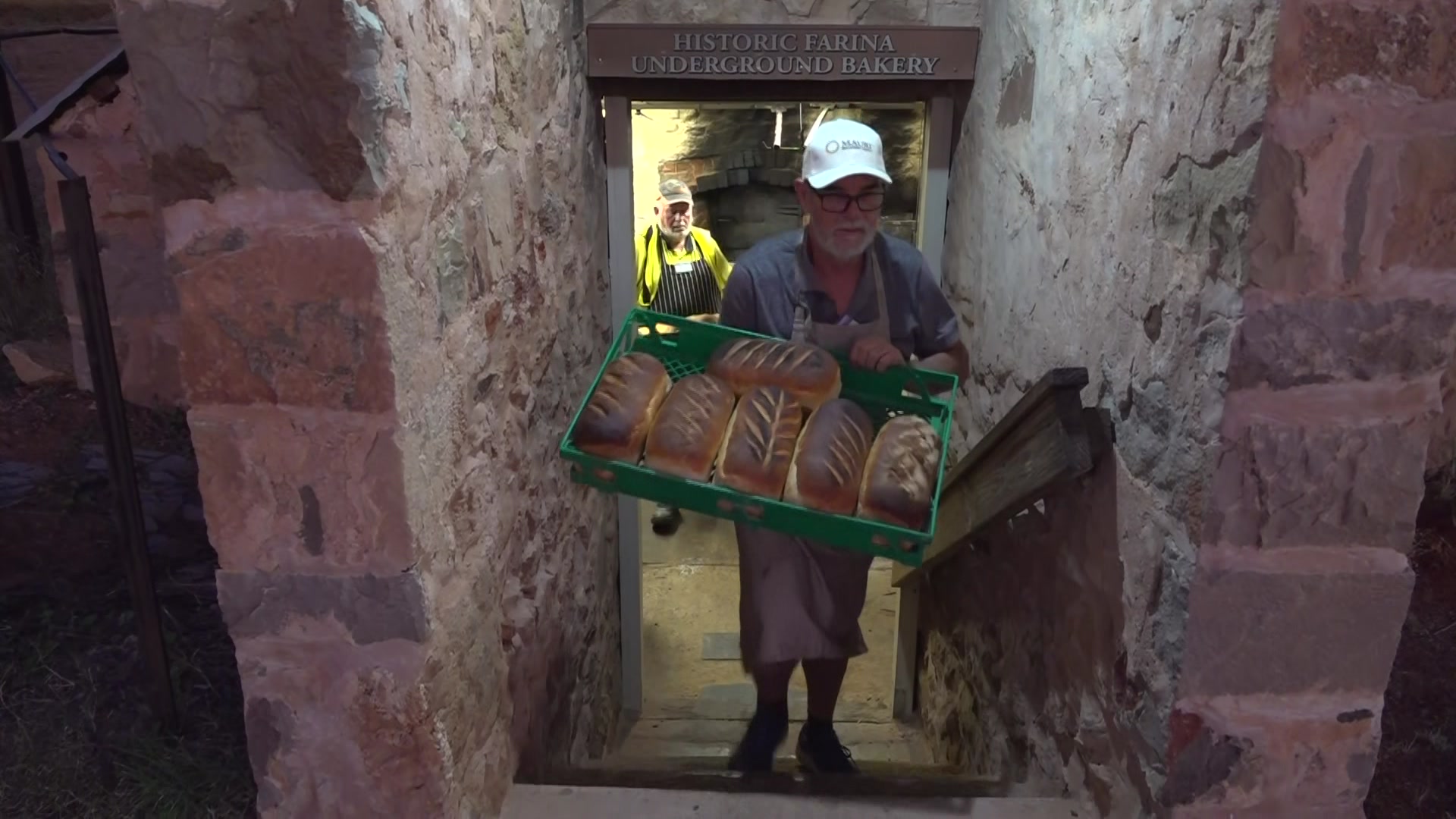 Two men carry trays of freshly baked bread loaves up steps from an underground baker.