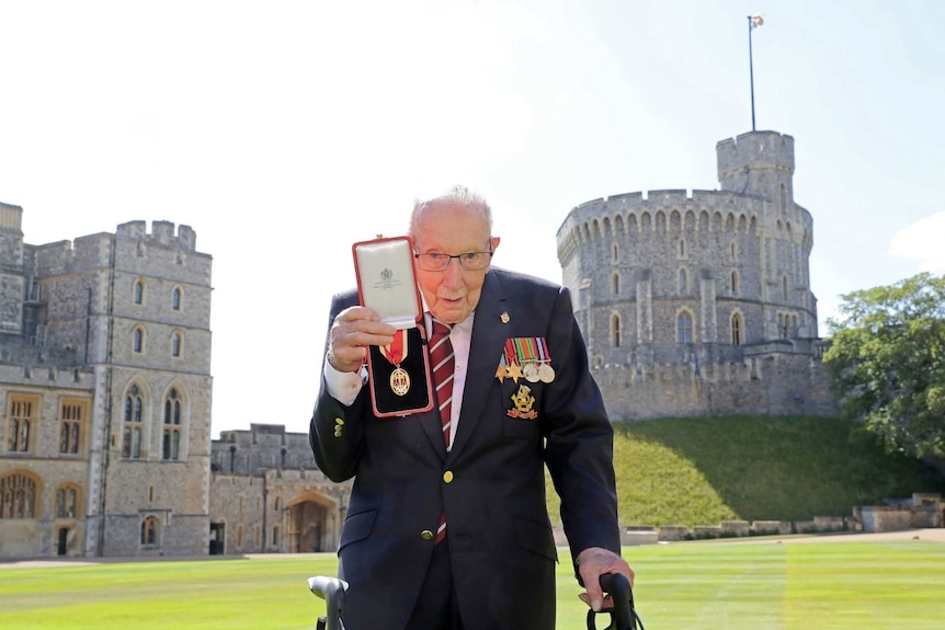 Sir Tom Moore wearing medals and a suit holds up his knighthood outside Windsor Castle
