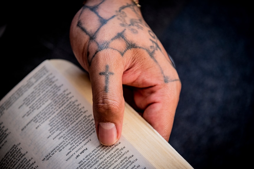 A tattooed hand, with a thumb with a cross tattoo, holds open a bible.