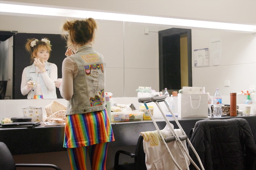 A woman wearing rainbow pants applies make-up in a dressing room mirror. 