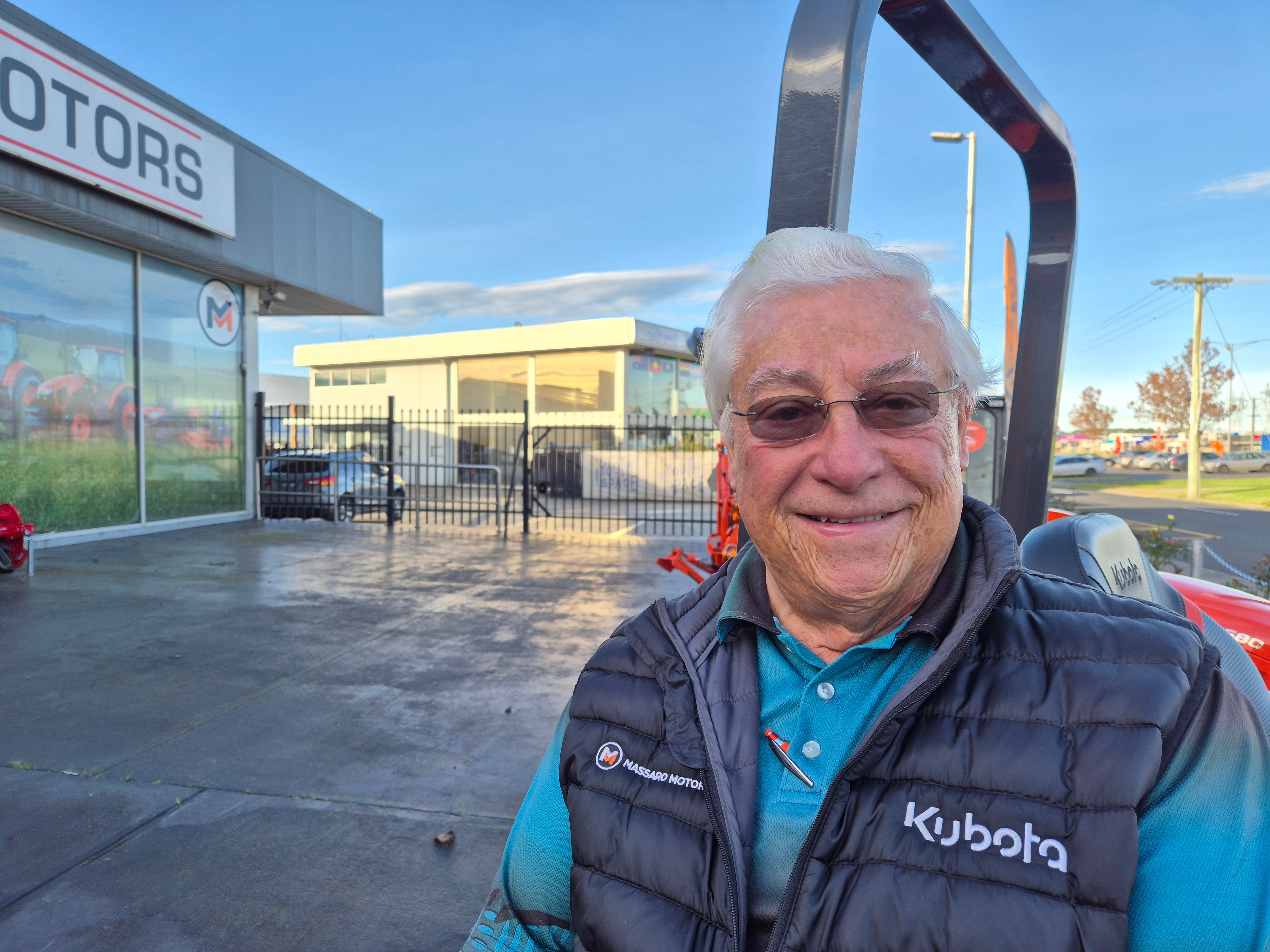 An elderly man in a puffer vest and blue tshirt outside Massaro Motors with Latrobe Youth Space behind him.