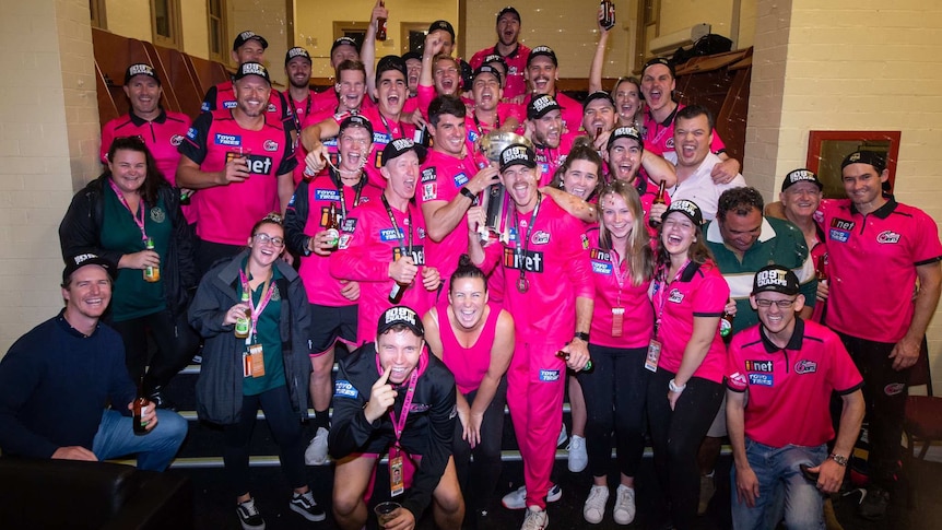 A group of men and women from the Sydney Sixers cheer and shout with bottles of beer in a changing room