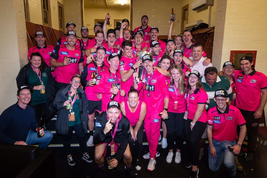 A group of men and women from the Sydney Sixers cheer and shout with bottles of beer in a changing room