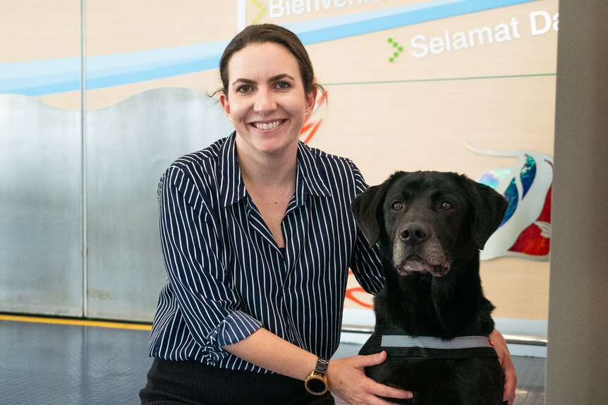 A woman wearing a striped shirt beside a black dog in front of metal automatic doors.