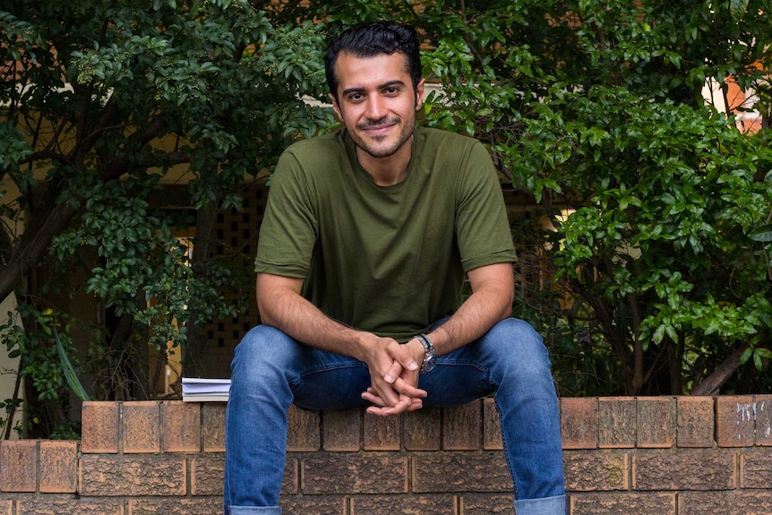 Shayan Askari sitting on a brick wall in front of hedge.