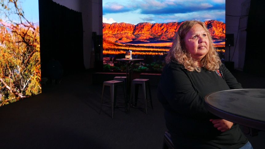 A woman sits at a chair and table looking into the distance. Behind her is a large photo of a rocky mountain range on a screen. 