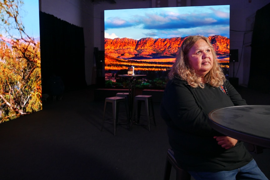 A woman sits at a chair and table looking into the distance. Behind her is a large photo of a rocky mountain range on a screen. 