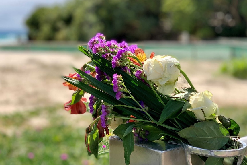 A close-up of a small bunch of purple, white and read flowers on ground with sea and trees behind.