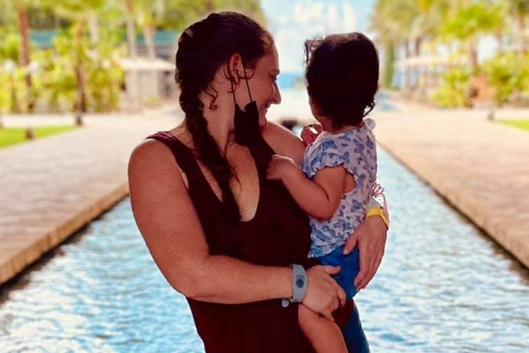 A mother holding her young daughter in her arms, standing in front of a water feature.