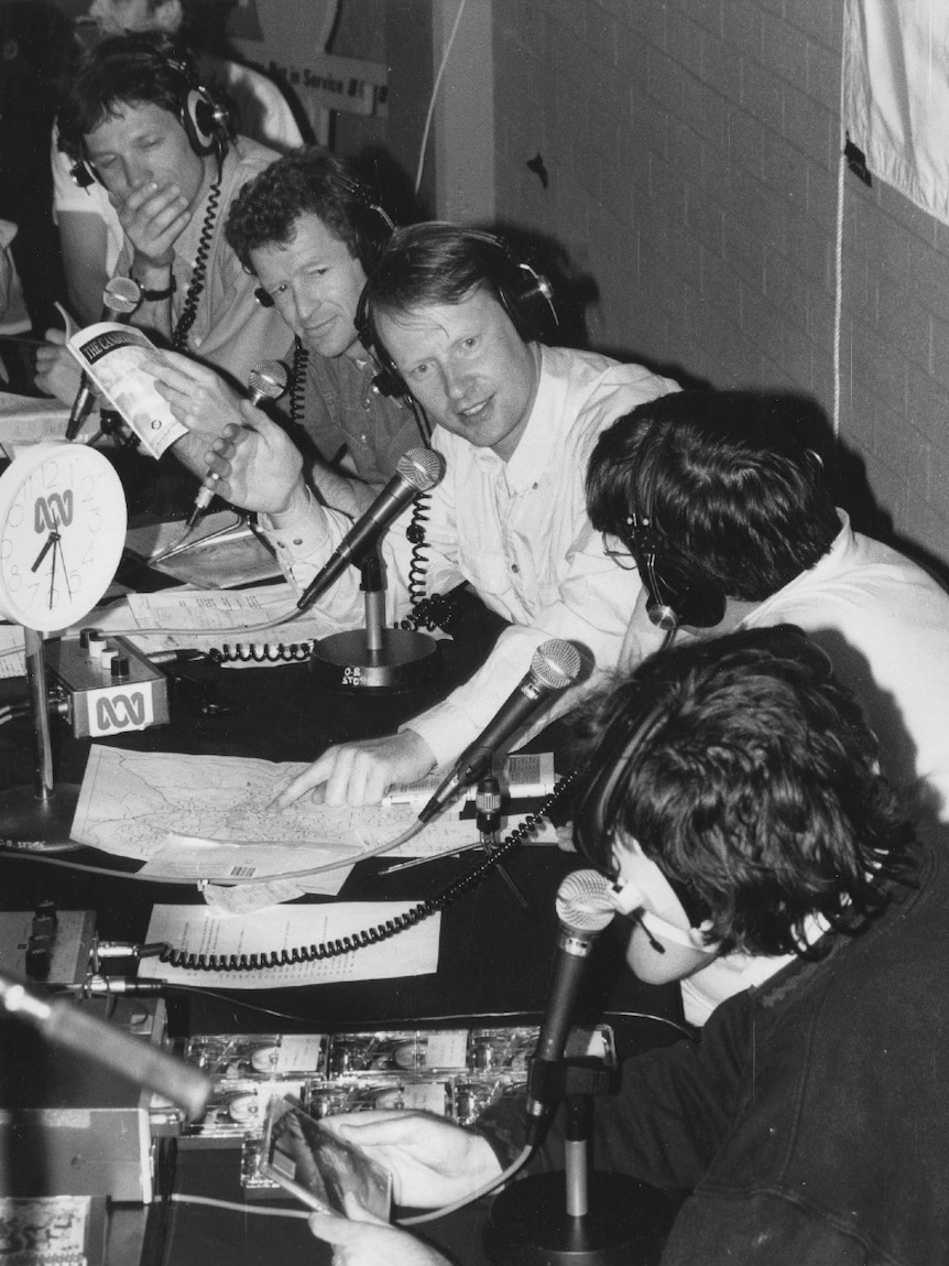 Black and white photo of five men lined up in front of microphones.