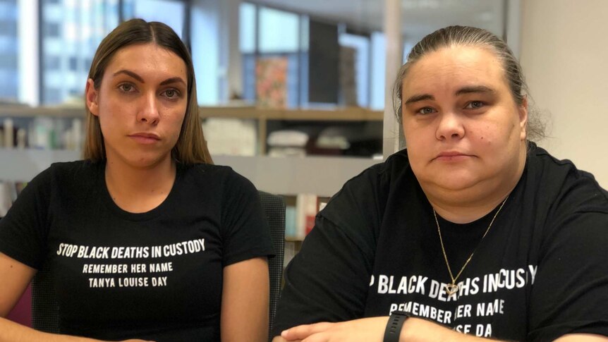 Two women wearing black t-shirts that say "Stop Black Deaths in Custody".