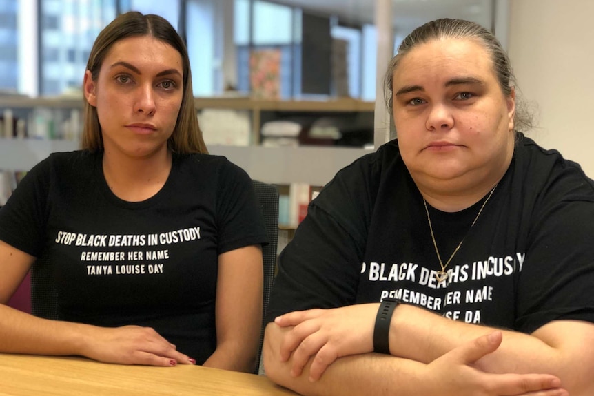 Two women wearing black t-shirts that say "Stop Black Deaths in Custody".