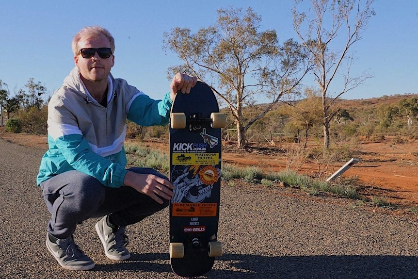 A man in his late 20s, fair-haired, wearing sunglasses, kneeling down and holding up a skateboard.