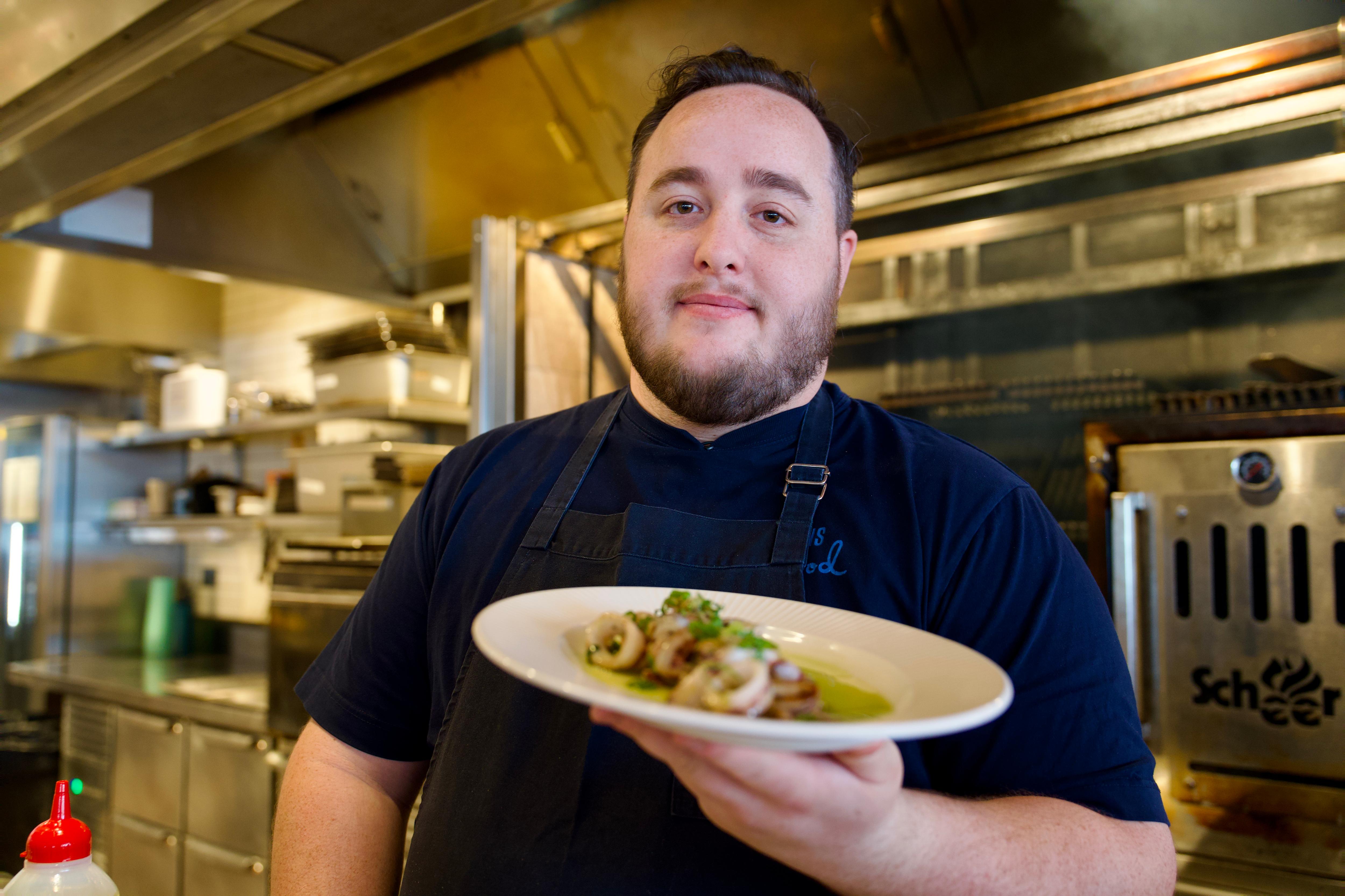 Luke Bourke stands in his kitchen holding a plate with a calamari dish, serving it up to the camera