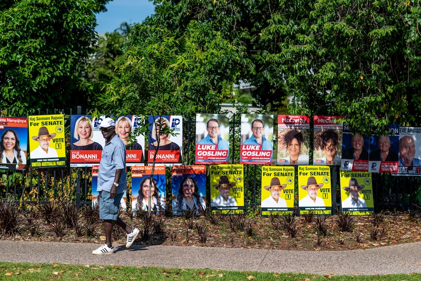 A man walks past a street corner covered in colourful election corflute posters.