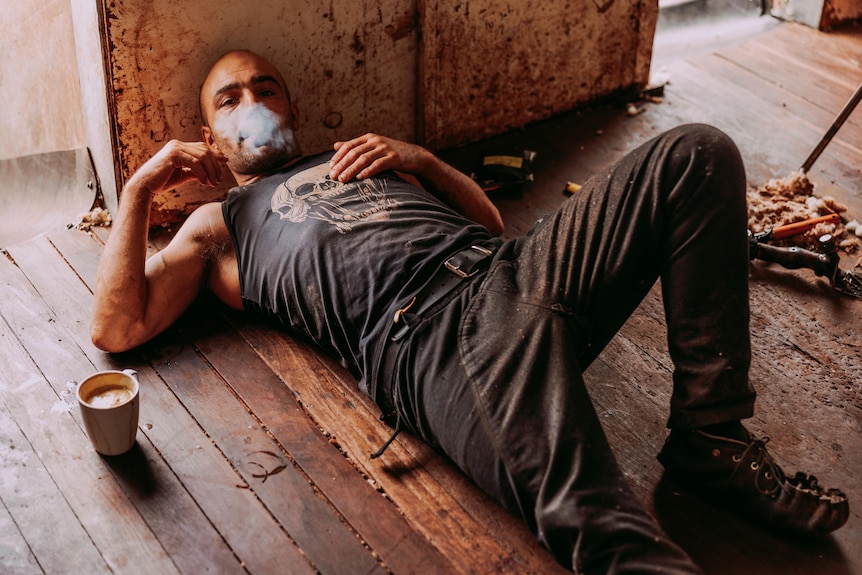 Man wearing a dark singlet and pants lies on the wooden floor of a shearing shed, with a mug of coffee next to him.