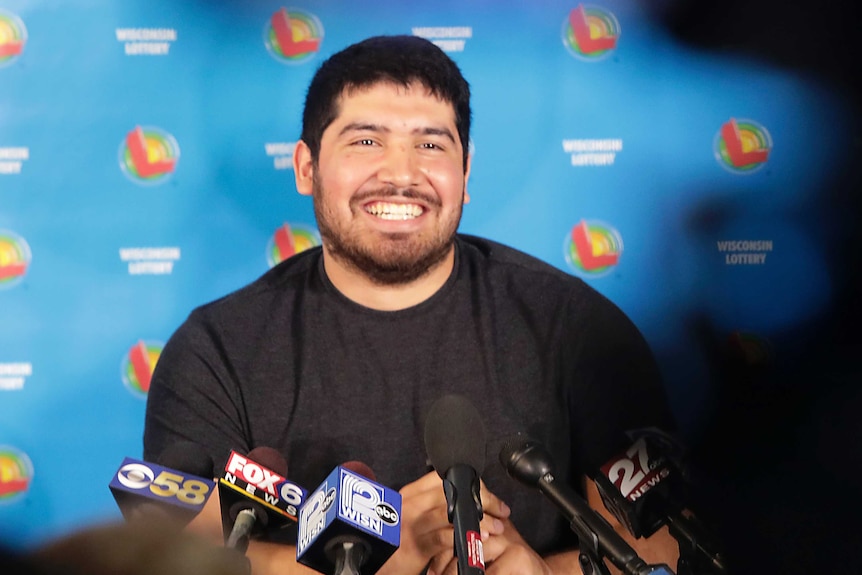 A man in a black shirt smiles at a press conference with microphones in front of him.