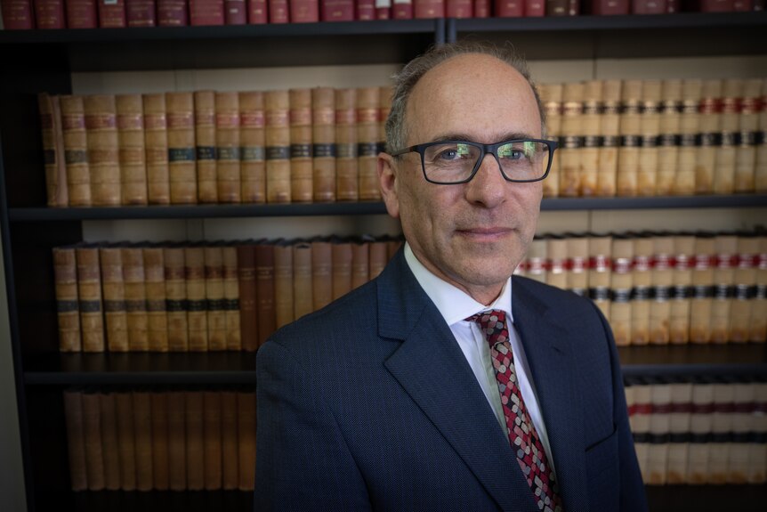 Roland Browne, wearing a suit and tie, stands in front of a bookshelf of law books