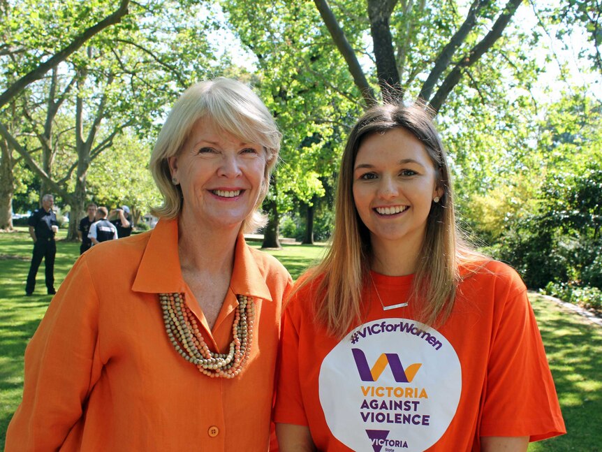 Two women wearing orange shirts are standing in park smiling at the camera.