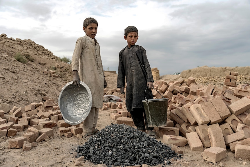 two boys stand next to a pile of bricks and black debris with a pan and bucket in their hands