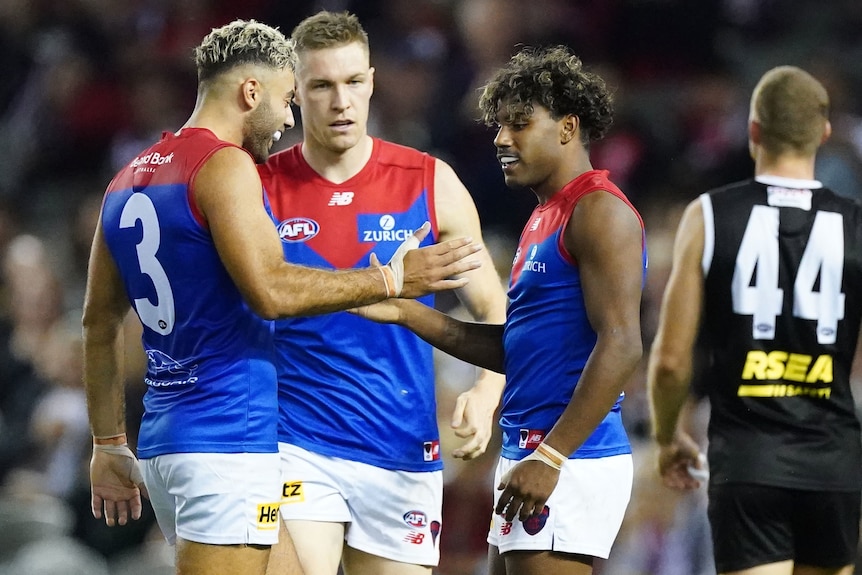 Two Melbourne Demons AFL players shake hands as a teammate looks on.