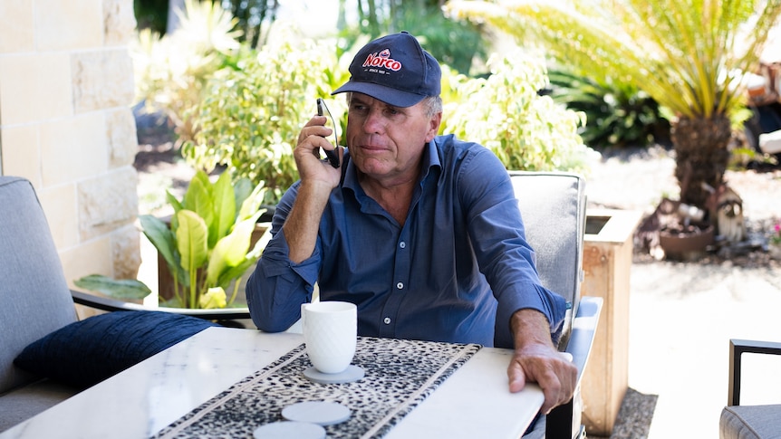 A man in a blue shirt and cap sits at a table listening to someone on his mobile phone.