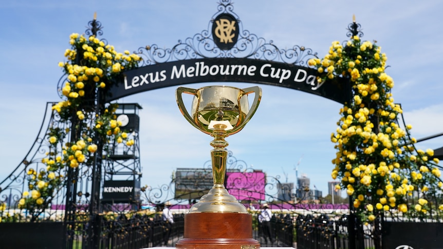 A solid gold trophy sits on a table in front of an archway with "Melbourne Cup Day" on it at the Flemington race