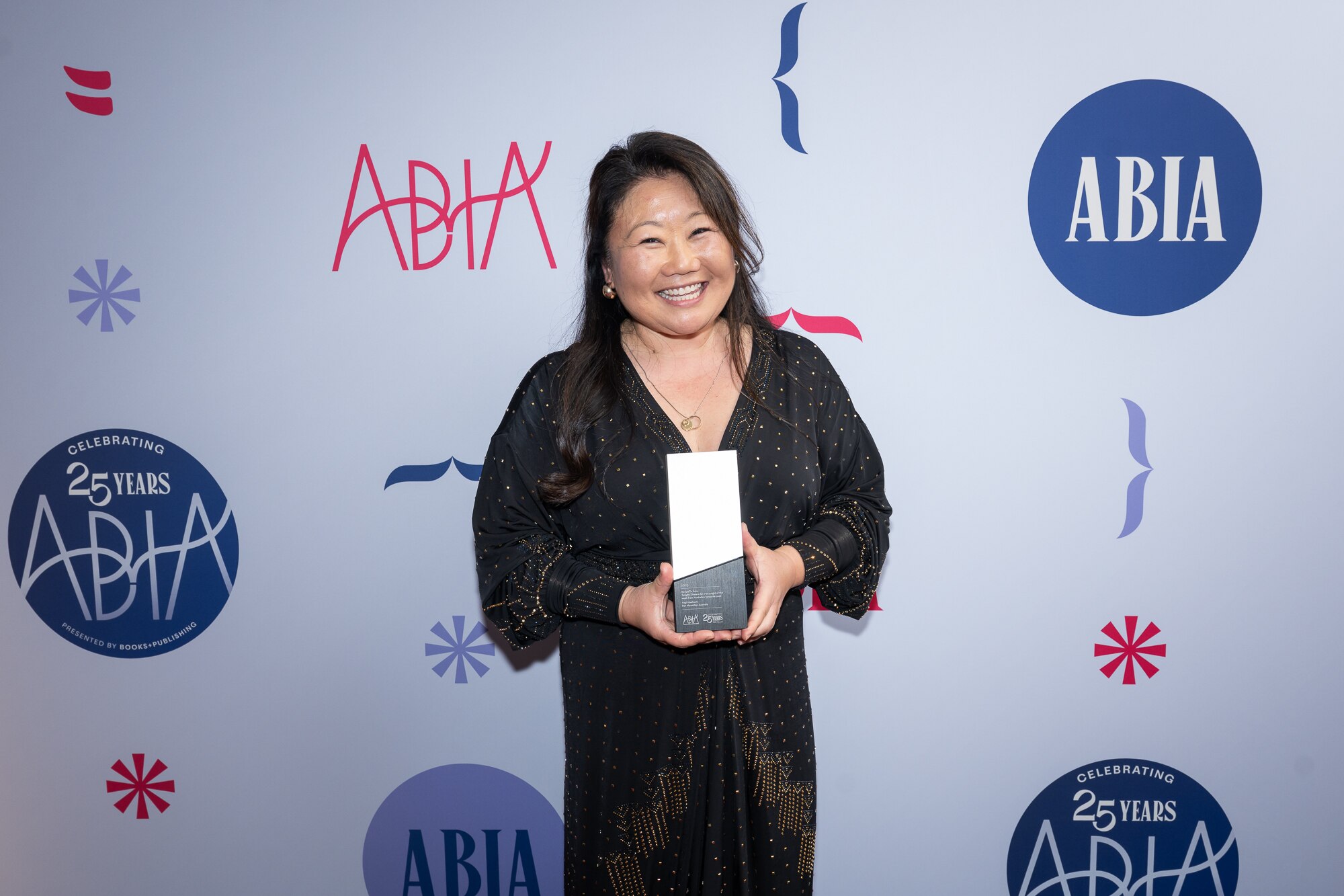 Nagi Maehashi, a Japanese Australian woman wearing a black dress, smiles brightly while holding a rectangle-shaped award.