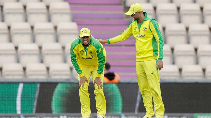 A cricketer grimaces in pain with his hands on his knees while a teammate pats him on the back.