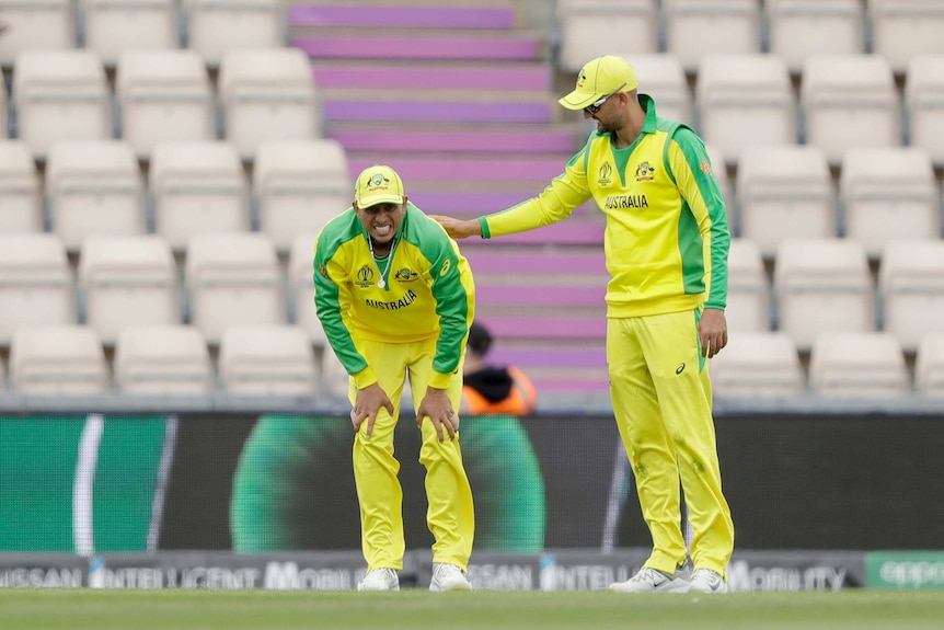 A cricketer grimaces in pain with his hands on his knees while a teammate pats him on the back.