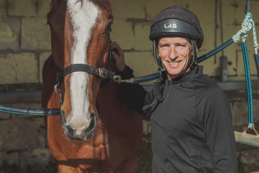 A man in a black riding helmet and long-sleeved black shirt smiles for a photo alongside a large thoroughbred horse.
