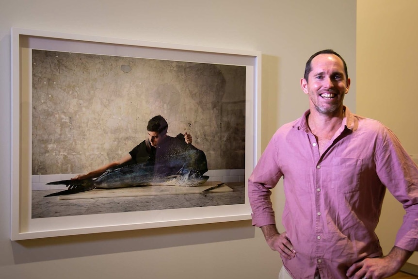 A man smiles while standing in front of a framer photograph of a man holding a fish.