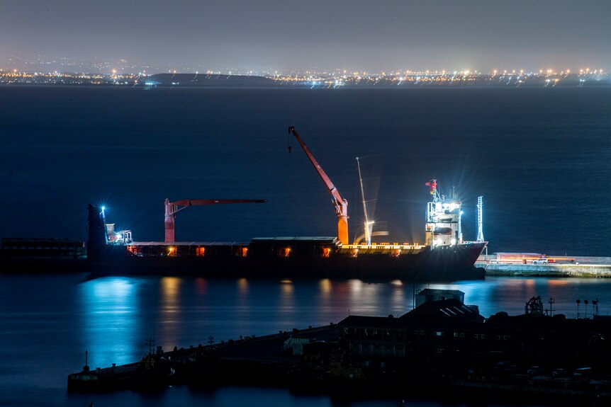 A Russian vessel is docked at night with a flat calm sea extending to city lights in the background.