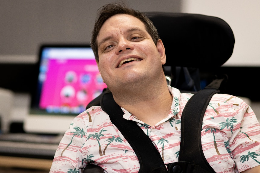 A smiling young man sitting on a wheelchair inside a room, with a lit-up computer screen in the background.