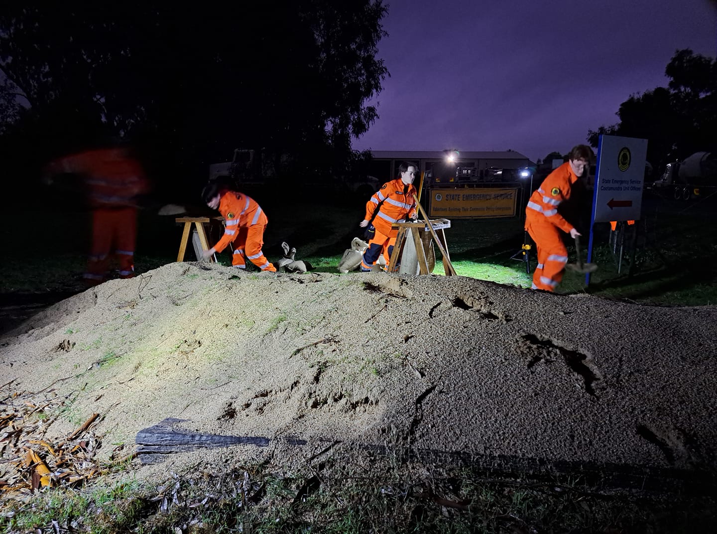 Three people in high-vis suits work to sandbag a verge at night.