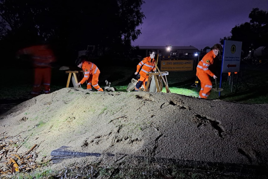 Three people in high-vis suits work to sandbag a verge at night.
