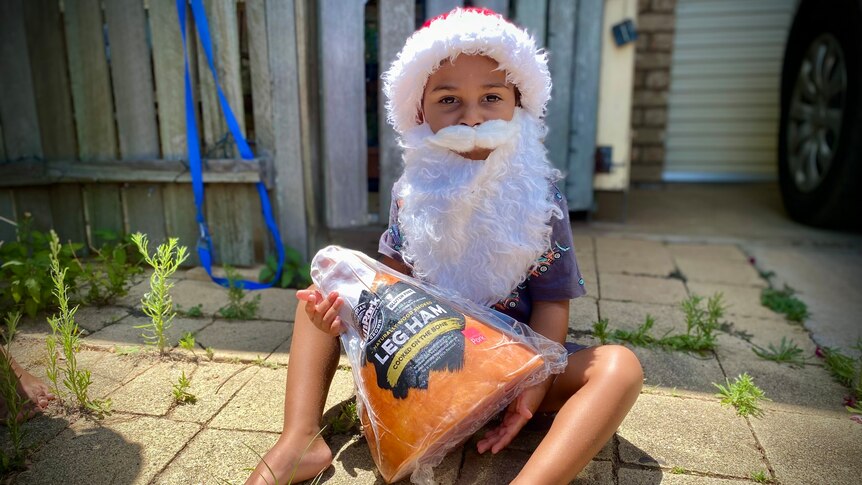 A child in a santa hat and beard holds a leg of ham half his own body size