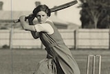 A girl in a tunic swings a cricket bat behind her head as another girl fields behind her