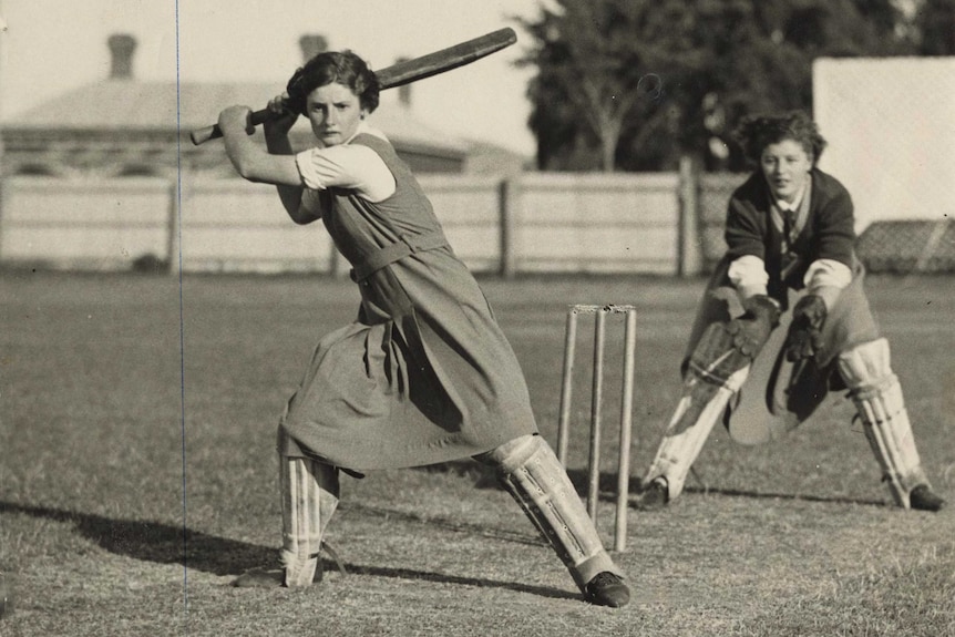 A girl in a tunic swings a cricket bat behind her head as another girl fields behind her
