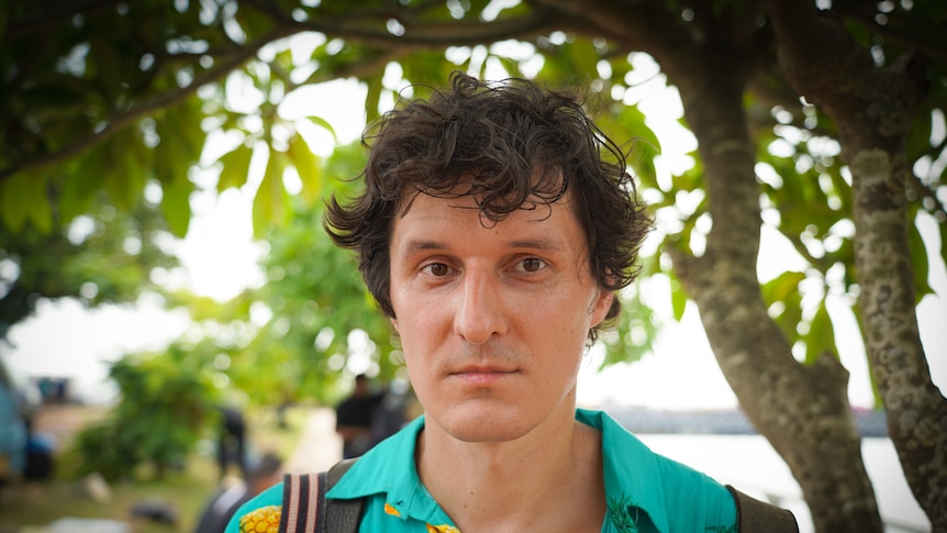 A young man with floppy brown hair and a loud pineapple-print shirt stands under a tropical tree