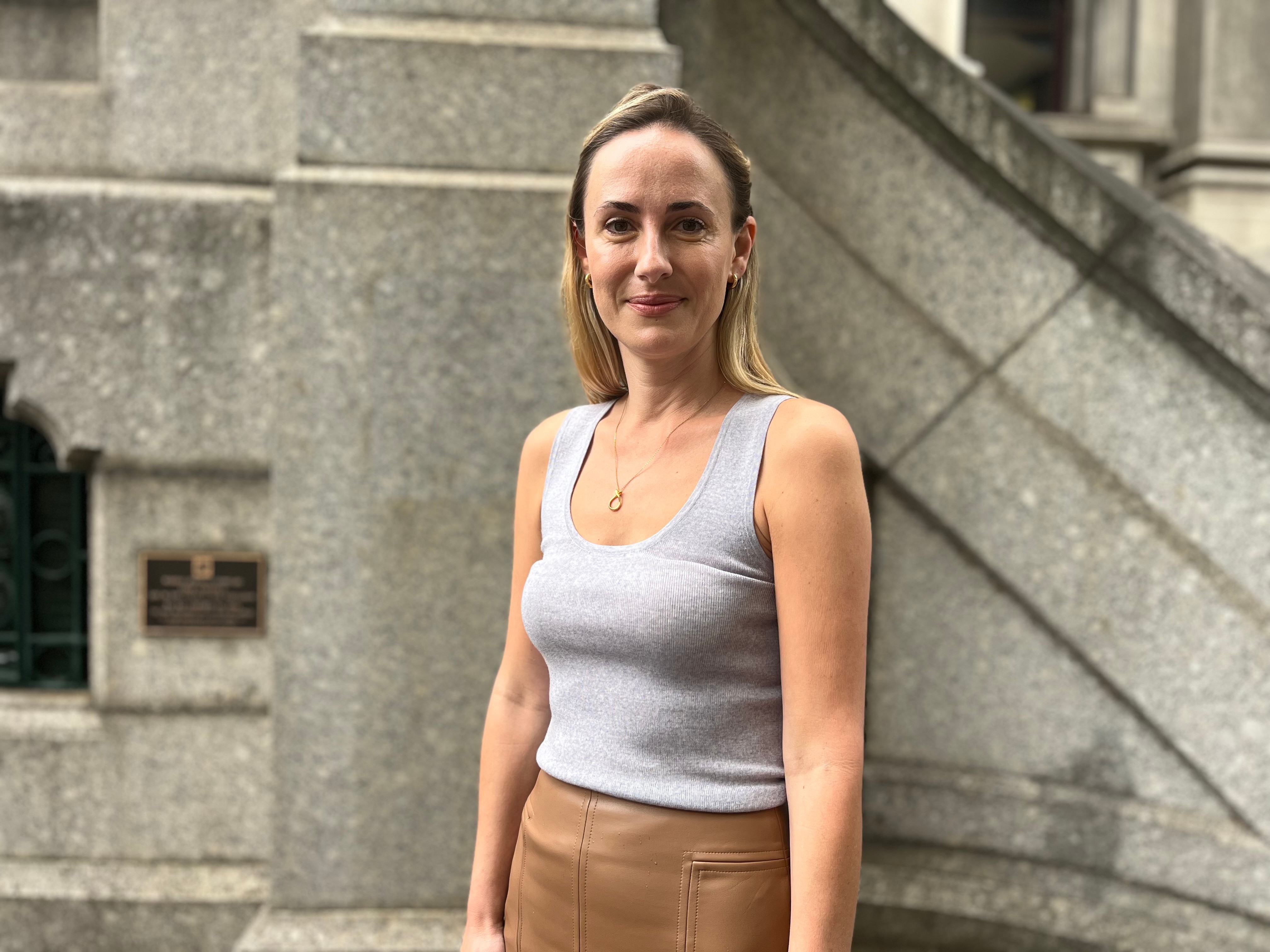 A woman with long blonde hair in a grey tank top and tan leather skirt smiles at the camera in front of a grey stone wall.