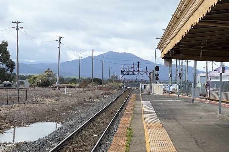 An empty train platform. Signals, a puddle, and a mountain are visible in the cloudy distance.